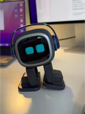 The image features a small, dark gray robot with a square head. The robot has blue rectangular eyes and is wearing blue headphones. It stands on a desk with two light-colored computer monitors in the background.