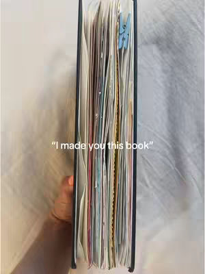 The cover image shows a book held vertically, with the edge of the pages facing the viewer. The pages are filled with colorful inserts and held together with a blue clip. A hand is visible, holding the book against a blurred, off-white background. White text overlay at the bottom of the image: "I made you this book".