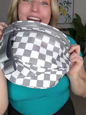 The cover image features a woman holding a checkered bag in front of her face, partially obscuring it. She is looking at the camera with her mouth open. She has blonde hair and is wearing a teal top. The background appears to be a living room setting, and includes a glimpse of decor like a picture frame and plants.