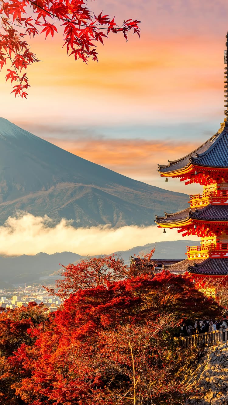 The image showcases a scenic view of a Japanese landscape with a focus on architectural and natural elements. In the foreground, vibrant red foliage of maple trees frames the lower portion and the left side of the frame. Emerging from behind the trees is a multi-tiered, reddish-orange pagoda with detailed architectural features, bells hanging from the eaves, and blue roofing. In the background, a large, majestic mountain (likely Mount Fuji) is visible, partially obscured by clouds. The sky has a warm gradient, transitioning from orange to blue. The overall composition blends natural beauty with cultural landmarks, creating a visually striking scene.
