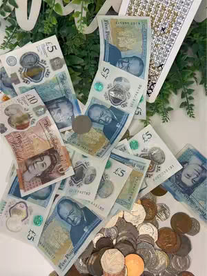 The image shows a pile of British banknotes and coins. Banknotes in denominations of £5 and £10 are spread out, with coins of various sizes and values forming a heap in the foreground. The arrangement suggests a display of money, possibly as a savings accomplishment.