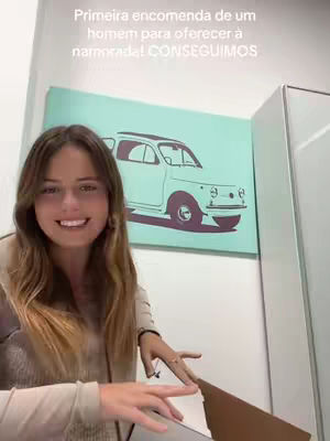 A smiling woman with long brown hair is captured in a well-lit room. She's wearing a beige top, and her left hand is resting on a partially opened cardboard box. Behind her, a light blue wall features an artwork of a white car. To her right, a white cabinet contrasts with the wall's color. Above the woman a hook text appears on the image.