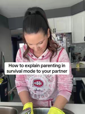 A woman is shown indoors at a kitchen sink, wearing a pink and white hoodie with a red and white logo, leaning over, with her arms in yellow gloves submerged, while looking down. The background features a white kitchen with various appliances. A white text overlay is present on the image.