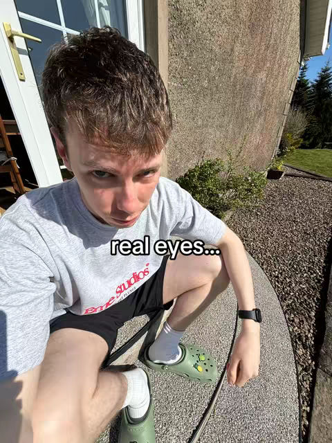 A selfie taken at a low angle, showing a young man sitting outside. He is wearing a gray t-shirt with a red and white logo, black shorts, and green sandals. The background includes a textured wall, a door, and greenery.