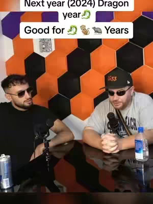 Two men are seated at a table, appearing to be in a studio setting. The background is a geometric wall pattern of orange, black, and purple hexagonals. The man on the left, wearing a black shirt and sunglasses, has a microphone in front of him, and a can and appears to be speaking. The man on the right, with a cap and sunglasses, also has a microphone, and a bottle of water is on the table.
