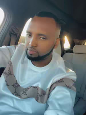 A man in a white shirt with a patterned design on the shoulders is sitting in a car. The man has short, dark hair and a trimmed beard. He appears to be looking at the camera. The interior of the car is visible with beige seating and the sunlight coming across the window.