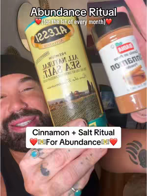 A close-up shot showing a man holding a salt container and a cinnamon container. The man has a beard, a nose ring, and tattoos on his arms. Text overlays on the image read 'Abundance Ritual' and 'Cinnamon + Salt Ritual For Abundance'.