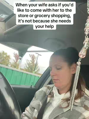 A woman is inside a car, presumably the driver's seat, with the window showing a view of outside with some green and brown colors. The woman is the center of focus.