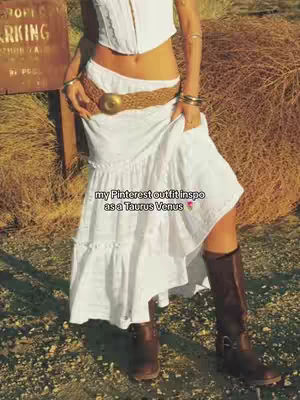 The image features a woman standing outdoors, dressed in a white top and a flowing white skirt. She wears brown boots. The background includes natural elements and a brown, weathered sign.