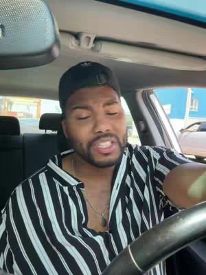A man is shown sitting in a car taking a selfie. He's wearing a black and white striped shirt and a black cap. The car's interior is visible, with a part of the car's seat and the steering wheel. The background includes a building's wall.