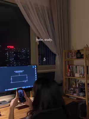 A person with dark hair sits at a desk in front of a window at night, holding a smartphone. A computer screen and a bookshelf are also visible. The text overlay includes the words "babe, study." and the number "5".