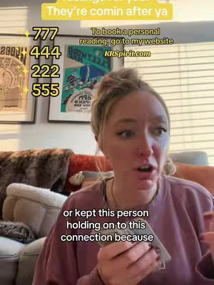 A woman with light skin and blonde hair, wearing a pink sweater, looks up and to the right with a surprised or concerned expression. Behind her is a couch, art pieces, and blinds.