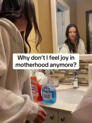 A woman is reflected in a mirror, with the primary view focused on cleaning supplies in the foreground of a bathroom. A text overlay reads, 'Why don't I feel joy in motherhood anymore?'