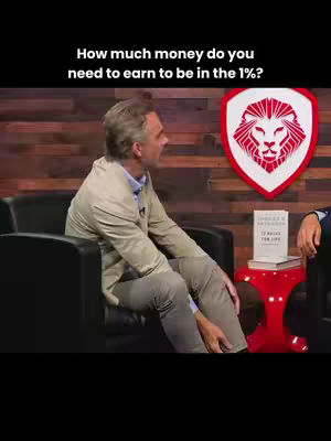 The image features a man seated in a chair, positioned to the left against a backdrop of wooden panels; he is engaged in what appears to be a discussion. To his right, there's a large, shield-like emblem featuring a lion's head. Below, a small red table supports a book and a partially visible hand. The overall mood seems conversational and informative, with warm lighting enhancing the setting.