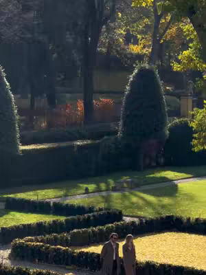 A couple walking in a park on a sunny day with lush green lawns and meticulously trimmed bushes. The scene is well-lit, casting shadows, and the colors are natural and vibrant.