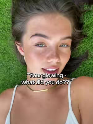 A close-up shot of a woman lying on a bed of green grass, facing upwards. Her face is centrally framed, with her eyes open and looking towards the camera. She has fair skin, visible eyebrows, and a slight smile, with text overlayed on the image.