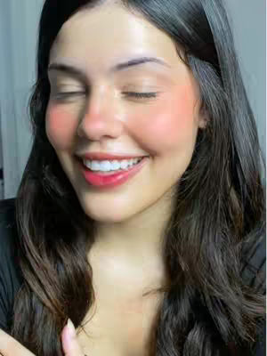 A close-up shot of a young woman with long, dark hair and a smiling face. Her eyes are closed. She has rosy cheeks and red lips. The background is out of focus.
