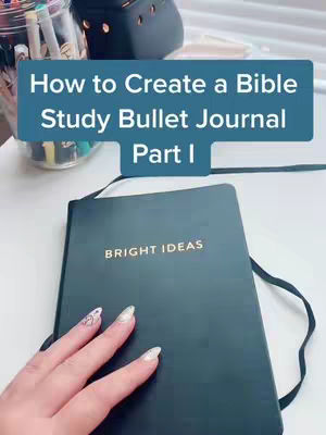 The image shows a black notebook on a white surface, with a hand partially covering the cover. The notebook is labeled 'BRIGHT IDEAS' in gold lettering. Above the notebook, a rectangular blue shape contains the text 'How to Create a Bible Study Bullet Journal Part I'. There are pens and other supplies in a jar visible in the background.