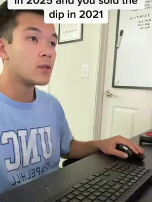 A young man is seated in front of a computer, looking at the screen with a thoughtful expression. He is wearing a light blue t-shirt with a dark blue graphic that says "UNC". The setting includes a white door, a white board or desk with notes on it, and a keyboard.