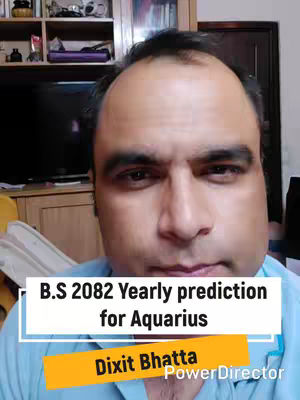 A man with short dark hair is centered in the frame, looking directly at the camera. The background features a blurred interior with wooden furniture and shelves. Overlay text in a white box reads "B.S 2082 Yearly prediction for Aquarius", with a smaller orange banner below containing the name "Dixit Bhatta" and the "PowerDirector" watermark.