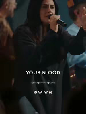 A woman with dark hair and a black hoodie is singing into a microphone. The image is taken from a live performance, with the background blurred. The text overlay has the words "YOUR BLOOD" in white font, and the name "@ Winnie" is at the bottom in a smaller font.