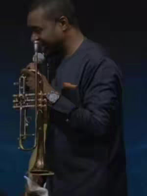 A man in a black long-sleeved shirt is playing a trumpet, the instrument held up to his mouth. The background is dark, suggesting a stage or performance setting. The lighting is dim.