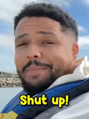 A man with short, dark curly hair and a beard is wearing a life jacket, with the sky and water in the background. The words "Shut up!" are overlaid in yellow text.