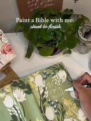 The image shows a close-up scene of someone painting a Bible. The Bible is open, revealing floral designs on a light green background. The person's hand holding a white pen is also visible. Additionally, there is a plant and several other items in the background.