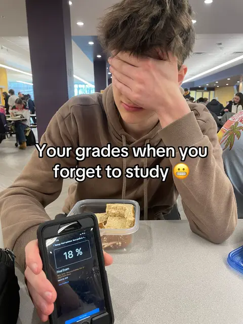 A young man covers his eyes with his hands, expressing disappointment. He's holding a phone displaying a low exam grade (18%). Beside him is a container of food. The setting appears to be a casual public space.