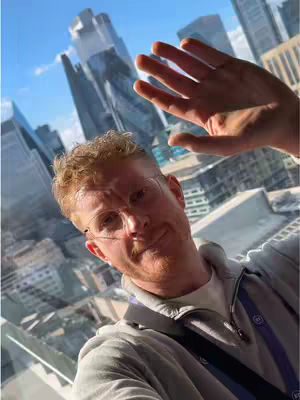 A selfie taken from a high angle with the creator in the mid-ground, in front of a large city panorama and a sky blue sky. The creator is wearing glasses and his hand is up as if blocking the sun. The pose suggests he is inside a building with large windows. The overall composition portrays a sense of achievement, possibly against the backdrop of an urban landscape or a business district.