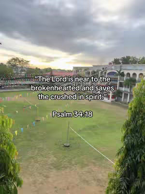 A wide outdoor shot during the day, dominated by a green field with colorful pennant flags in the background. In the distance, there are buildings and trees under a partly cloudy sky. Text including a bible verse is overlaid on the center-top of the image.