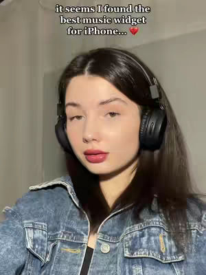 A young woman with dark hair wearing a denim jacket and black headphones is looking at the camera. The background is a blurred indoor setting. Text overlayed above her head reads, "it seems I found the best music widget for iPhone..." with a small heart emoji.