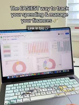 A laptop displaying a spreadsheet with charts and graphs is the central focus. Above the laptop, text reads 'The EASIEST way to track your spending & manage your finances' in bold white font, with a small money bag icon next to it. Below the text is an overlay with 'Link in bio' in a box. The keyboard cover has colorful stickers. The background appears to partially show a white desk and a window with blinds.