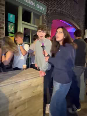 A woman interviews a young man on the street holding a microphone. Other people are in the background.