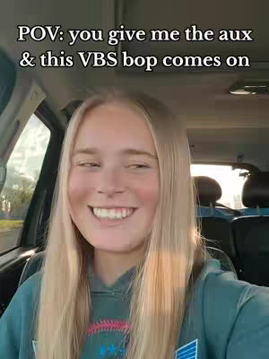 A selfie from inside a car. A young woman with long blonde hair is smiling widely at the camera. Text overlayed on the image reads, "POV: you give me the aux & this VBS bop comes on."
