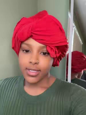 A woman with brown skin and a nose ring wears a dark green shirt and a large, vibrant red head wrap that covers her hair. She is facing the camera, with a neutral expression and slightly parted lips as if she's talking. A mirror is visible behind her to her right, reflecting a similar view of her head covering.