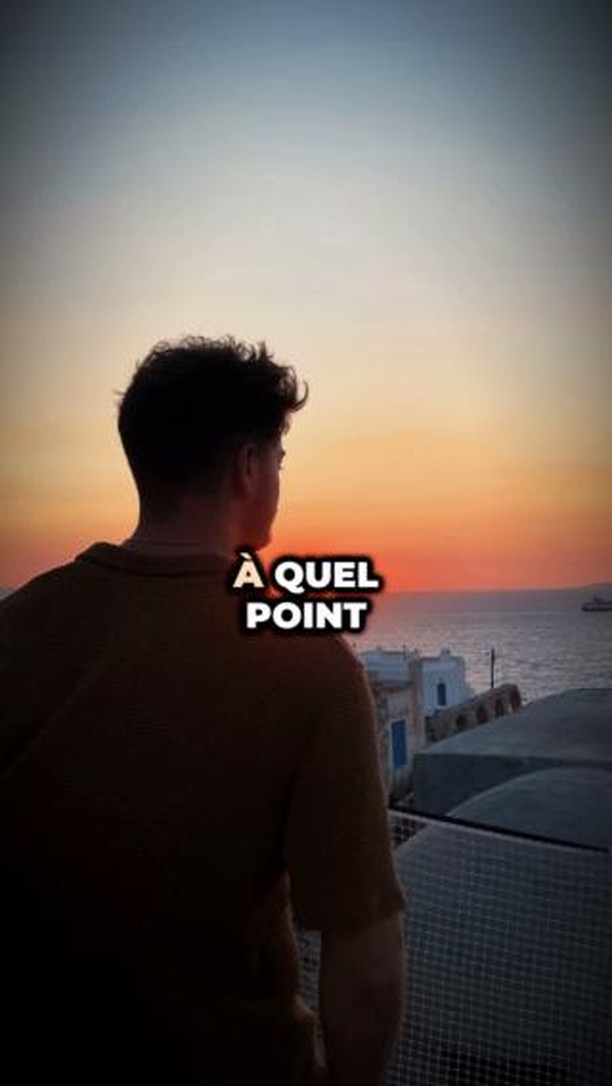 A man stands on what appears to be a rooftop, gazing towards a sunset over the sea. The sky transitions from orange to blue, with the sun just below the horizon. In the foreground, a portion of the rooftop is visible, with buildings and the sea in the distance. The text overlay is bold and white, providing context to the scene.