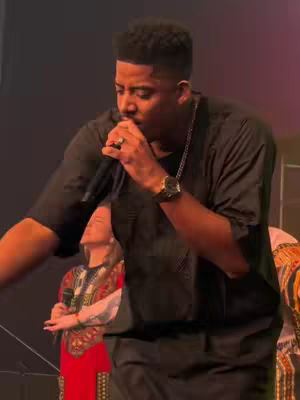 A man is shown singing into a microphone on a stage. He is wearing a black shirt and a gold watch. Two women in the background are singing along.