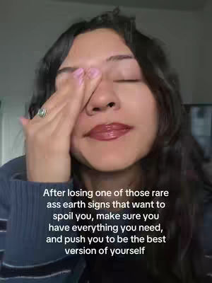 A close-up shot of a person with long, dark wavy hair, obscuring their right eye with their left hand. Their other eye is closed. Their lips are glossy and red and they appear to be wearing a dark blue sweater. Text overlayed at the bottom of the image.