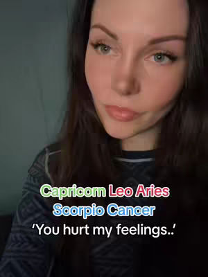 A selfie-style shot featuring a woman with long brown hair and fair skin gazing forward. The background is a muted bluish-gray shade. A colored overlay adds text: "Capricorn Leo Aries/Scorpio Cancer/‘You hurt my feelings..’"