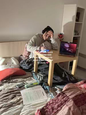 A young woman is sitting on a bed, leaning towards a small desk holding a laptop and a notebook, with a pillow and stuffed animal behind her. The bed is covered with a patterned comforter. Various study materials are spread out on the bed. The lighting appears to be ambient, with no harsh shadows.