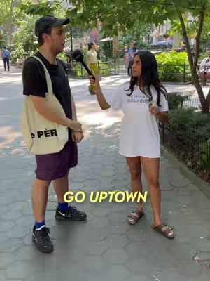 Two people are standing and talking in a park setting. One person holds a microphone and appears to be interviewing the other. The text 'GO UPTOWN' is overlaid near the bottom.