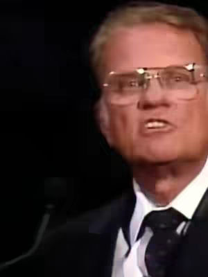 A medium shot shows a man with short, light hair, wearing glasses and a dark suit with a black tie. He's looking slightly to the side, with his mouth open as if speaking or singing. The background is a plain, dark color.