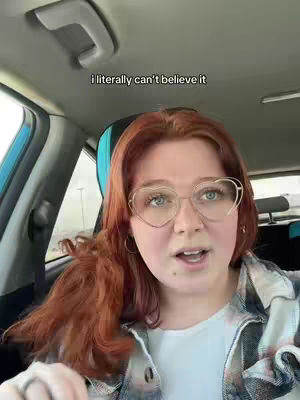 A woman with long red hair and heart-shaped glasses looks directly at the camera. She is seated inside a car, with the door and headrest visible in the background. The car's interior and the exterior environment outside are visible.