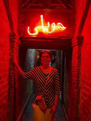 A woman stands in a dimly lit, red-hued alleyway. The walls are made of red bricks, and a neon sign with Arabic script hangs above. The woman is wearing a striped shirt and carrying a purse.