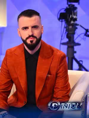 A man with short dark hair, a neatly trimmed beard, and a serious expression sits in a white armchair. He's wearing an orange blazer over a black shirt. The background is a blue wall, and part of a camera and lights are visible on the right. At the bottom of the image, a logo that says "Opinion" is seen.