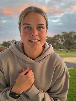 A young woman with blonde hair wearing a tan hoodie stands in front of a slightly blurred natural background. The sky has a mix of orange and blue hues, suggesting either sunrise or sunset. Her hands are clasped in front of her chest, conveying a sense of warmth or sincerity. She's smiling and appears directly into the camera, with a relaxed expression. A watch is visible on her left wrist. The overall image has a soft, natural aesthetic.