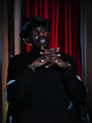 A man in a black outfit and hat sits with his hands clasped, against a red curtain backdrop.
