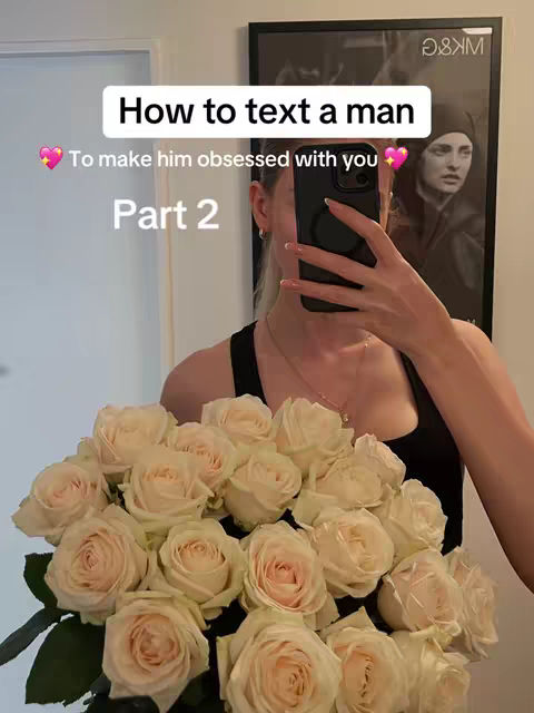 A selfie-style image with a woman holding a phone in front of her face, obscuring most of it. She has a bouquet of light pink roses in her arms, and appears to be in a room with a painting and a white door frame visible in the background.