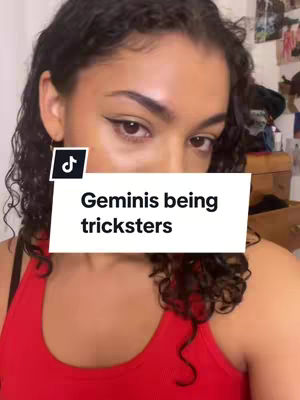 A selfie-style video cover featuring a young woman with dark, curly hair and makeup, looking directly at the camera. She is wearing a red top. An overlay on the image displays the text: 'Geminis being tricksters'. The background is a blurred interior suggesting a room setting.